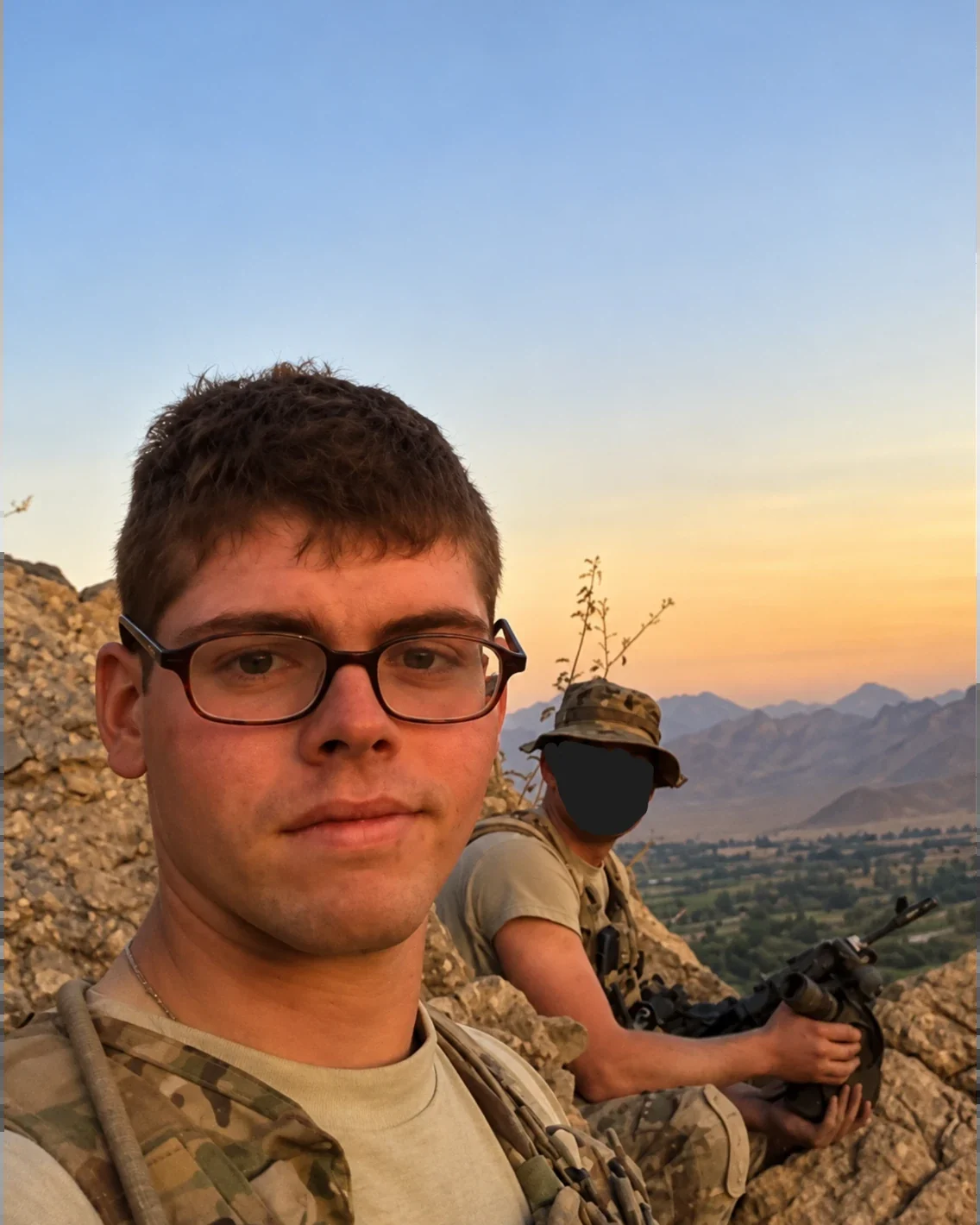 Jacob Ross on a ridgeline in Afghanistan at dusk, with a fellow soldier on watch and a valley below.