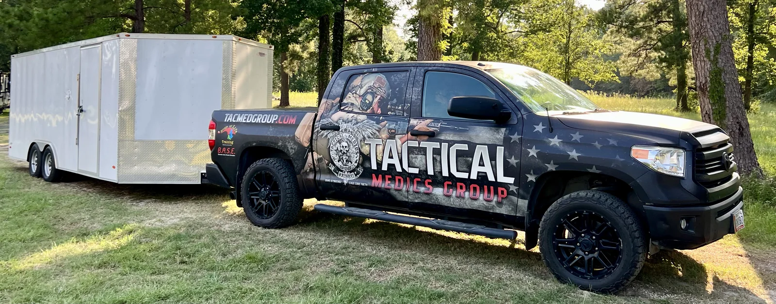 A Tactical Medics Group pickup truck towing an enclosed white equipment trailer, parked on grass at a wooded training site.